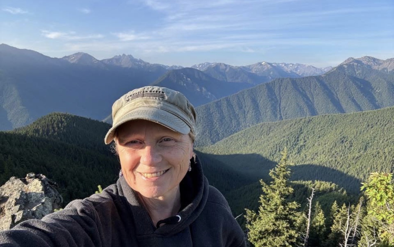 Jean Ball smiling at Deer Park overlook with Olympic Mountains in the background
